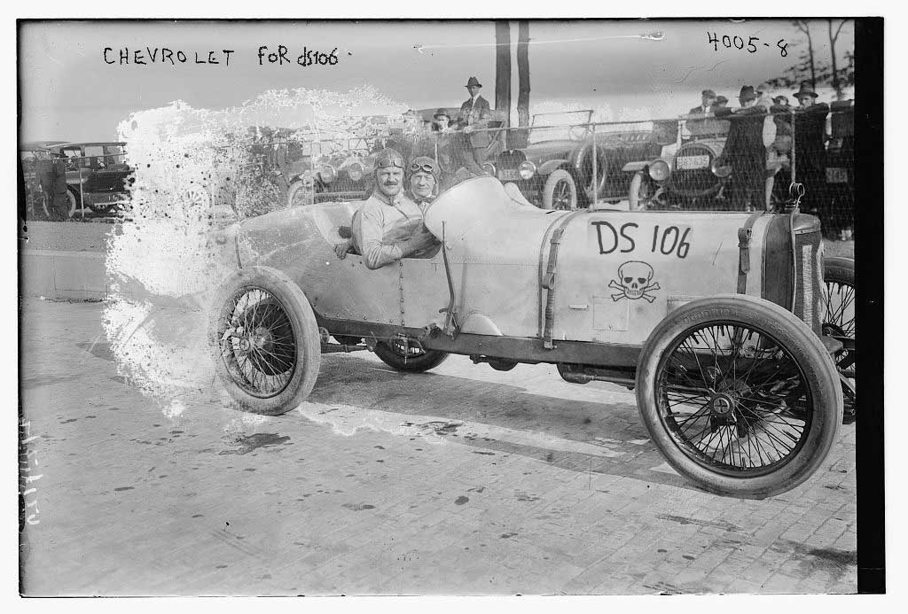 Antique and somewhat damaged photo of 2 men in racing hats and goggles sitting in a 1915 style car. On the side of the hood, someone has pasted some kind of skull and cross bones logo with DS106 on it