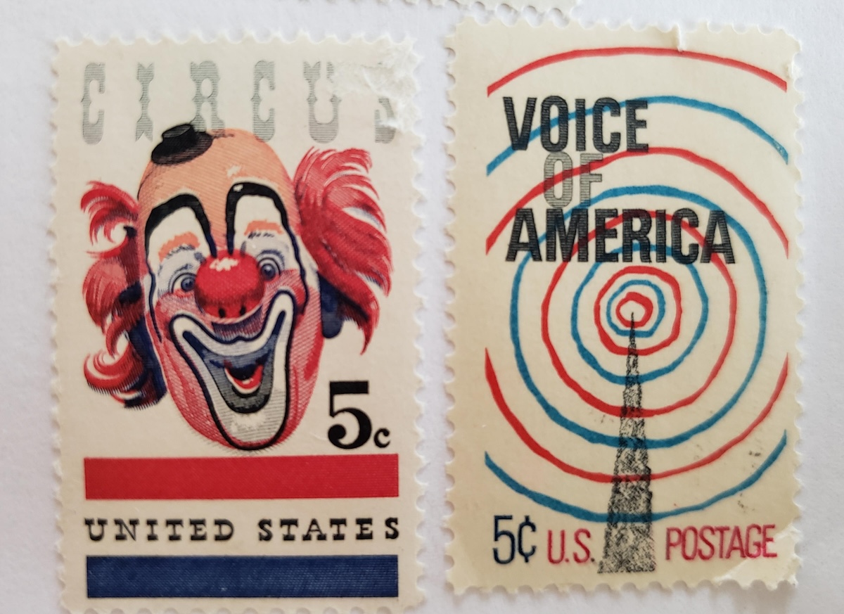 2 old U.S. 5 cent stamps. The one on the left has CIRCUS at the top faintly in grey, and a red, white and black clown . He has a big red nose, tiny black hat, red hair, and the usual clownface makeup. The one on the right has VOICE OF AMERICA at the top and a radio tower with green and red circles symbolizing airwaves.